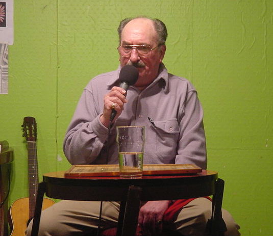 Gordon Cragg seated at bingo board with pint glass of water talking into the mike and eyeing the glass with deep suspicion